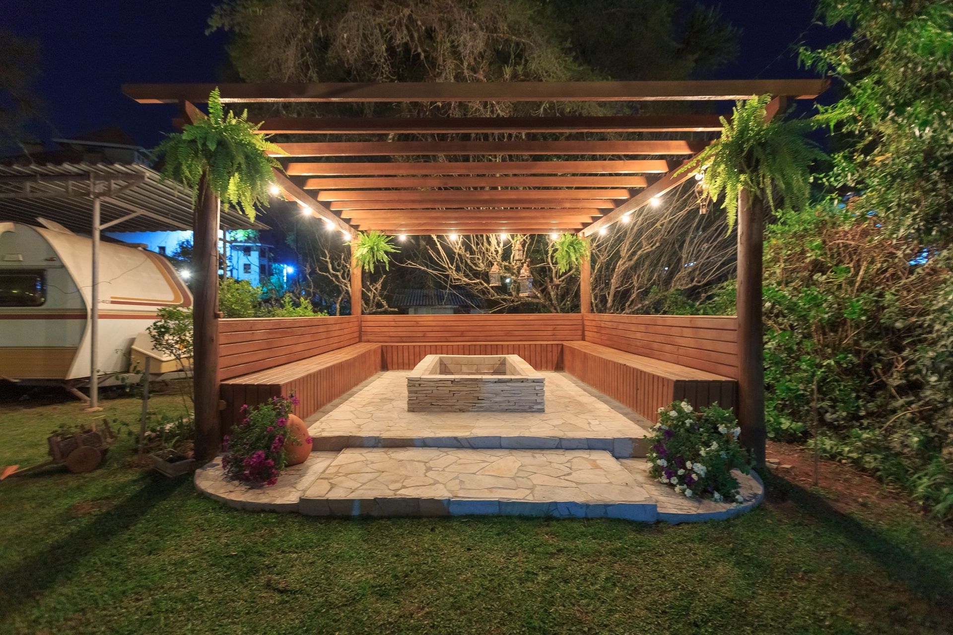 Illuminated wooden gazebo with fireplace and benches in garden setting at night.
